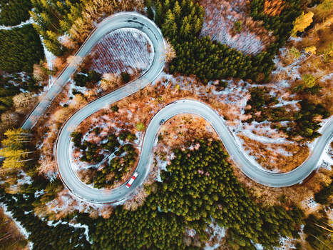Aerial view of a winding serpentine road cutting through a dense forest with patches of snow and autumn foliage in Czechia.