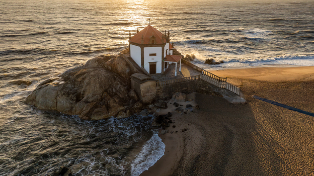 Aerial view of Capela do Senhor da Pedra on a rocky outcrop surrounded by crashing waves and golden sunset light Miramar Porto Portugal.