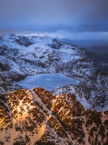 Aerial view of the sun kissed mountain peaks contrast with the icy lake reflecting the twilight sky creating a serene and majestic landscape Narvik Nordland Norway.