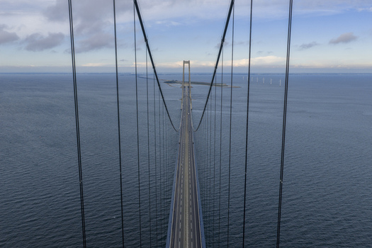 Aerial view of the Great Belt Bridge with its massive suspension cables stretching over the blue sea towards the horizon and distant wind turbines in Denmark. Aerial view of the Great Belt Bridge with its massive suspension cables stretching over the blue sea towards the horizon and distant wind turbines in Denmark.