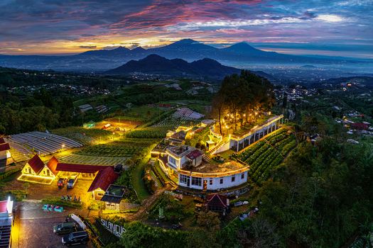 Aerial view of Silancur Highland with illuminated buildings and terraced fields against mountains under an orange sunset sky Magelang Regency Central Java Indonesia.