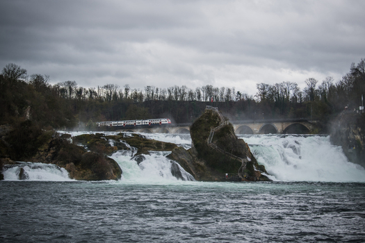 View of the Rhine Falls where the powerful cascade crashes around a central rock formation and a train gracefully crosses the bridge Schaffhausen Switzerland. View of the Rhine Falls where the powerful cascade crashes around a central rock formation and a train gracefully crosses the bridge Schaffhausen Switzerland.
