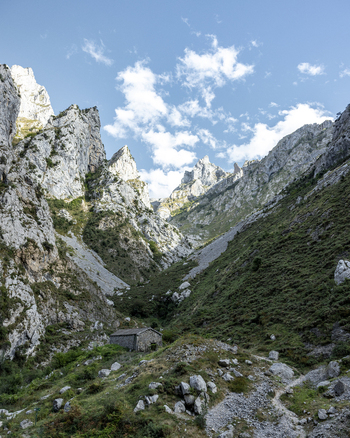 View of rugged towering peaks frame a lush valley where a small stone structure nestles amidst vibrant greenery under a vast sky Picos de Europa Asturias Spain. View of rugged towering peaks frame a lush valley where a small stone structure nestles amidst vibrant greenery under a vast sky Picos de Europa Asturias Spain.
