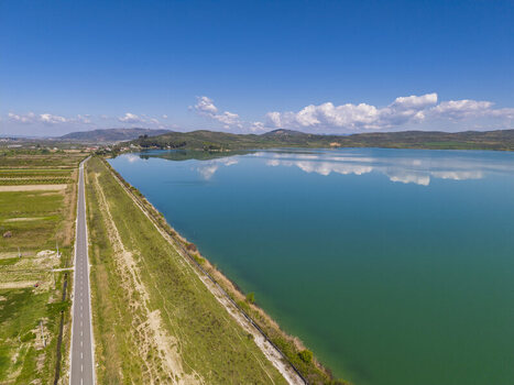 Aerial view of Liqeni Thanes serene reservoir with turquoise water reflecting the blue sky bordered by a long asphalt road and green agricultural fields in Lushnje Fier County Albania. Aerial view of Liqeni Thanes serene reservoir with turquoise water reflecting the blue sky bordered by a long asphalt road and green agricultural fields in Lushnje Fier County Albania.