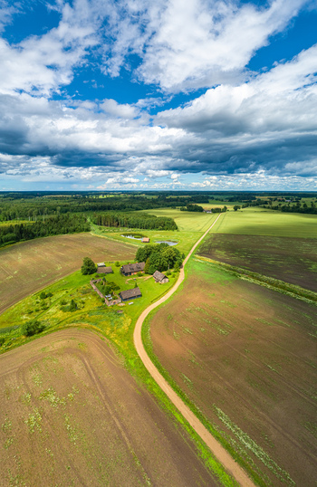 Aerial view of a rural farmstead surrounded by agricultural fields meadows and a dirt road under a blue sky with clouds in Latvia. Aerial view of a rural farmstead surrounded by agricultural fields meadows and a dirt road under a blue sky with clouds in Latvia.