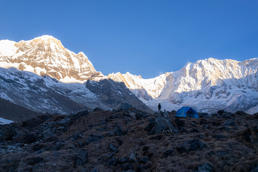 Aerial view of a blue tent and a hiker on a rocky slope beneath the snow capped peaks of the Annapurna mountain range in Nepal.