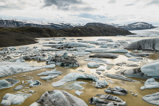 Aerial view of Vatnajokull glacier lagoon with floating icebergs in the glacial water surrounded by rugged mountains and a cloudy sky in Iceland.