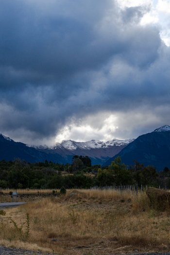 View of Parque Nacional Los Alerces with snow capped mountains under dramatic dark clouds and golden grass in the foreground in Futaleufu Department Chubut Province Argentina. View of Parque Nacional Los Alerces with snow capped mountains under dramatic dark clouds and golden grass in the foreground in Futaleufu Department Chubut Province Argentina.