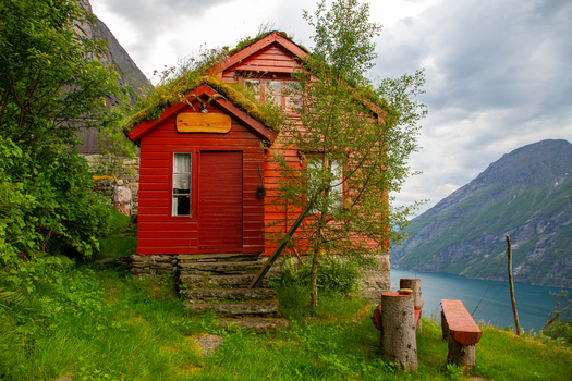 View of Smage mountain farm a traditional red wooden house with a grass roof overlooking the fjord surrounded by lush green grass and steep mountains in Geiranger More og Romsdal Norway.