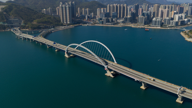Aerial view of the Cross Bay Link bridge with its iconic white arch spanning over the blue water towards the city skyline and green hills in Hong Kong Hong Kong.