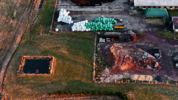 Aerial view of a rural farmstead featuring stacks of white and green hay bales a small pond and piles of earth under the warm light of the golden hour in Latvia. Aerial view of a rural farmstead featuring stacks of white and green hay bales a small pond and piles of earth under the warm light of the golden hour in Latvia.