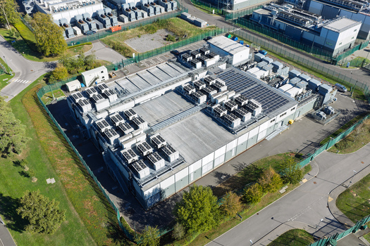 Aerial view of a data center facility featuring numerous rooftop HVAC units and solar panels enclosed by a perimeter fence in the United Kingdom.