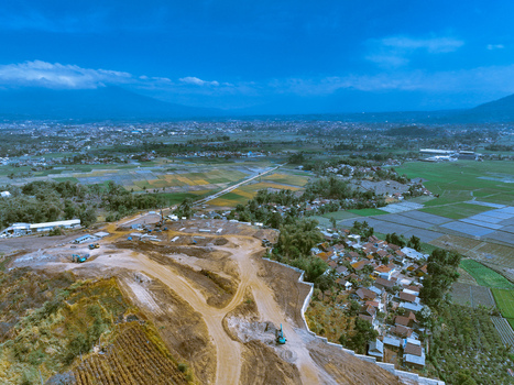 Aerial view of a construction site with heavy machinery and excavated earth surrounded by lush green rice fields and a residential area in Garut West Java Indonesia. Aerial view of a construction site with heavy machinery and excavated earth surrounded by lush green rice fields and a residential area in Garut West Java Indonesia.