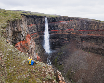 Aerial view of Hengifoss waterfall with iconic red clay layers and basalt cliffs featuring hikers sitting on the edge of the canyon in Egilsstadir Fljotsdalshreppur Iceland. Aerial view of Hengifoss waterfall with iconic red clay layers and basalt cliffs featuring hikers sitting on the edge of the canyon in Egilsstadir Fljotsdalshreppur Iceland.