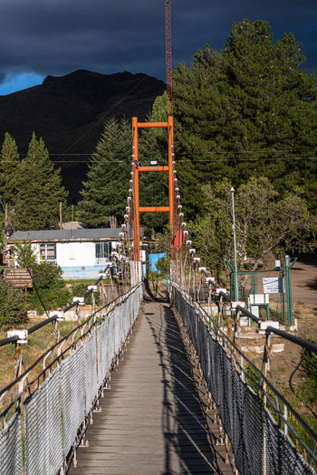 View of the suspension bridge at Villa Llanquin with orange metal towers and wire mesh railings set against pine trees and mountains in Cipolletti Rio Negro Argentina. View of the suspension bridge at Villa Llanquin with orange metal towers and wire mesh railings set against pine trees and mountains in Cipolletti Rio Negro Argentina.