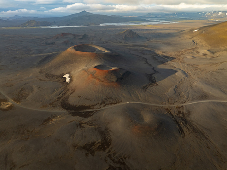 Aerial view of Landmannalaugar featuring volcanic craters with dark soil and reddish rims winding gravel road and distant mountains in Rangarthing ytra Iceland. Aerial view of Landmannalaugar featuring volcanic craters with dark soil and reddish rims winding gravel road and distant mountains in Rangarthing ytra Iceland.