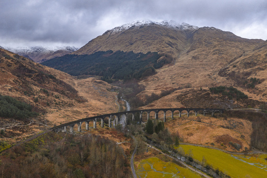 Aerial view of Glenfinnan Viaduct curving through the rugged Highlands landscape with snow capped mountains and a winding river under a cloudy sky Glenfinnan Scotland United Kingdom.