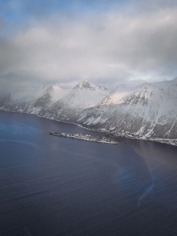 Aerial view of the stark contrast between the snow covered mountain peaks and the dark still waters surrounding a small village Fjordgård Troms Norway. Aerial view of the stark contrast between the snow covered mountain peaks and the dark still waters surrounding a small village Fjordgård Troms Norway.