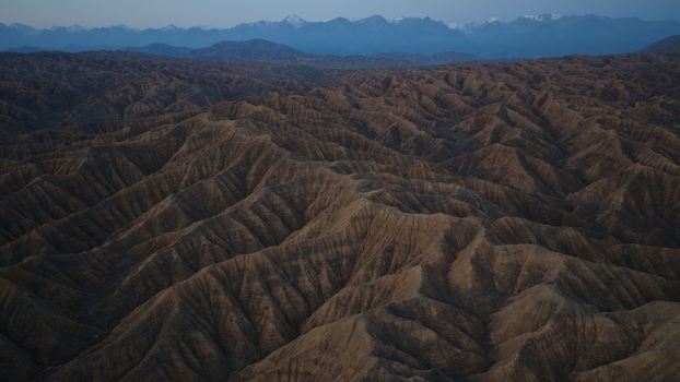 Aerial view of the rugged eroded rock formations of Skazka Canyon with snow capped mountains in the distance under a twilight sky in Issyk Kul Region Kyrgyzstan. Aerial view of the rugged eroded rock formations of Skazka Canyon with snow capped mountains in the distance under a twilight sky in Issyk Kul Region Kyrgyzstan.
