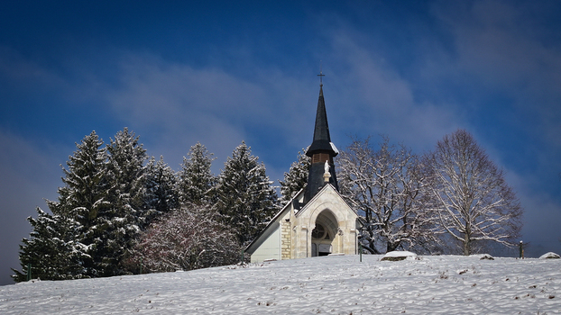 Aerial view of a small stone chapel on a snow covered hill with evergreen trees under a blue sky in Vesancy Auvergne Rhone Alpes France. Aerial view of a small stone chapel on a snow covered hill with evergreen trees under a blue sky in Vesancy Auvergne Rhone Alpes France.