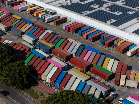 Aerial view of a large logistics shipping container yard filled with colorful cargo containers and a warehouse building with solar panels on the roof in Carson California United States.