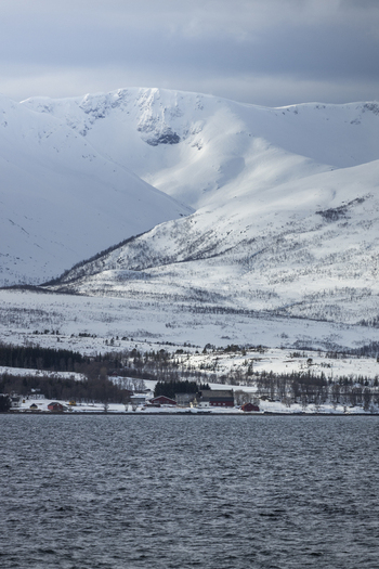 View of snow capped mountains and a small coastal settlement along the fjord under a cloudy sky Tromso Troms Norway.