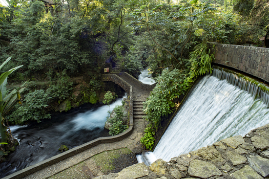 View of Parque Nacional Barranca del Cupatitzio featuring a stone bridge over a flowing river and a cascading waterfall surrounded by lush forest in Uruapan Michoacan Mexico. View of Parque Nacional Barranca del Cupatitzio featuring a stone bridge over a flowing river and a cascading waterfall surrounded by lush forest in Uruapan Michoacan Mexico.