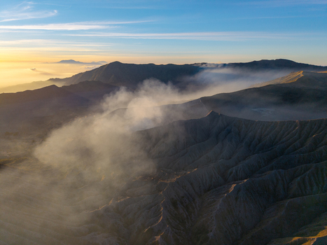 Aerial view of Mount Bromo sunrise with volcanic smoke rising over the rugged caldera and surrounding mountain peaks in the golden morning light in Tosari East Java Indonesia. Aerial view of Mount Bromo sunrise with volcanic smoke rising over the rugged caldera and surrounding mountain peaks in the golden morning light in Tosari East Java Indonesia.