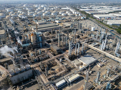 Aerial view of an industrial oil refinery featuring numerous storage tanks processing units piping networks and steam plumes rising from the facility in Carson California United States.
