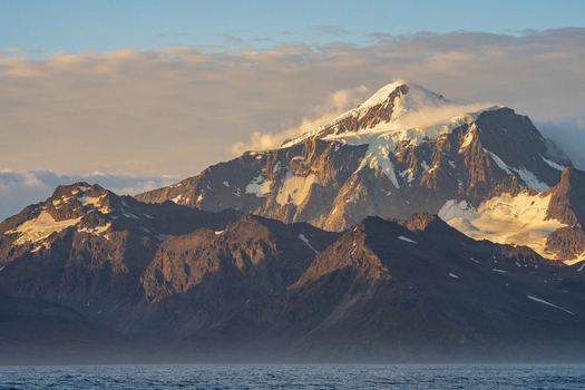 View of snow capped mountain peaks near Grytviken illuminated by golden hour light with rugged slopes and calm sea Grytviken South Georgia and the South Sandwich Islands.
