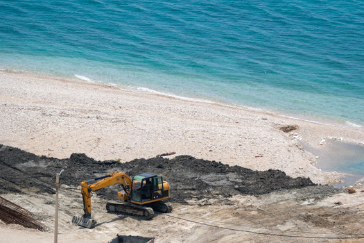 View of a yellow excavator working on a construction site next to a pebble beach and the turquoise sea in Vlore County Albania. View of a yellow excavator working on a construction site next to a pebble beach and the turquoise sea in Vlore County Albania.