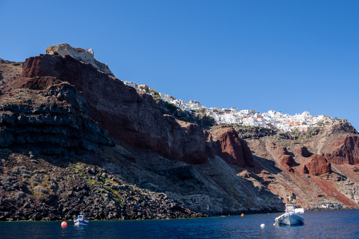 View of the steep volcanic cliffs of Santorini with white houses of Fira perched on the ridge above the blue Aegean Sea with small boats Fira Greece. View of the steep volcanic cliffs of Santorini with white houses of Fira perched on the ridge above the blue Aegean Sea with small boats Fira Greece.