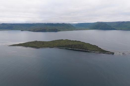 Aerial view of a small green island surrounded by the calm waters of a bay with distant mountains under a cloudy sky in Unalaska Alaska United States.