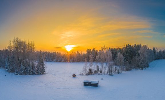Aerial view of a snow covered landscape with a small wooden cabin and a barrel sauna surrounded by frost covered trees under a vibrant golden sunset in Latvia. Aerial view of a snow covered landscape with a small wooden cabin and a barrel sauna surrounded by frost covered trees under a vibrant golden sunset in Latvia.