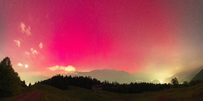 View of the aurora borealis glowing pink over the mountain peaks silhouetted forests and a small building in Valtellina Sondrio Lombardy Italy. View of the aurora borealis glowing pink over the mountain peaks silhouetted forests and a small building in Valtellina Sondrio Lombardy Italy.