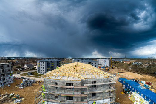 Aerial view of a building under construction with wooden roof trusses and scaffolding workers in high visibility vests on site set against a dramatic dark stormy sky in Latvia. Aerial view of a building under construction with wooden roof trusses and scaffolding workers in high visibility vests on site set against a dramatic dark stormy sky in Latvia.