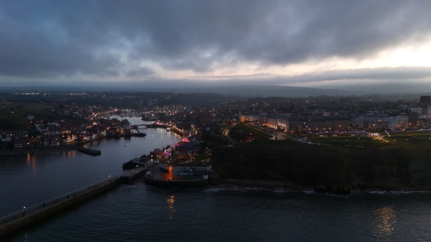 Aerial view of a coastal town harbor at twilight with illuminated buildings a long pier and harbor lights reflecting on the water under a cloudy sky United Kingdom.