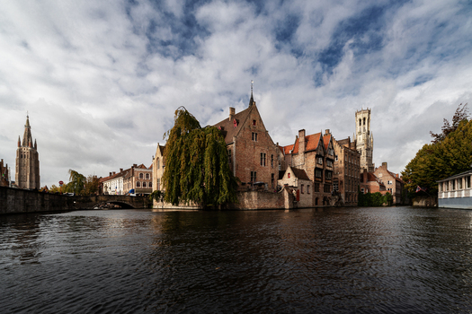 View of historic brick buildings along the canal with the Belfry and Church of Our Lady under a cloudy sky in Bruges Flanders Belgium.