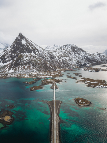 Aerial view of a road stretching across turquoise waters towards snow capped mountains Flakstadbruene Nordland Norway. Aerial view of a road stretching across turquoise waters towards snow capped mountains Flakstadbruene Nordland Norway.
