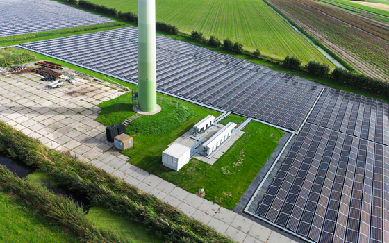 Aerial view of a wind turbine base surrounded by extensive solar panel arrays in a rural landscape with green fields and irrigation ditches in t Zand North Holland Netherlands. Aerial view of a wind turbine base surrounded by extensive solar panel arrays in a rural landscape with green fields and irrigation ditches in t Zand North Holland Netherlands.