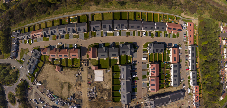 Aerial view of a modern residential housing development featuring rows of houses with solar panels private gardens and parked cars along a curved road near a construction site.