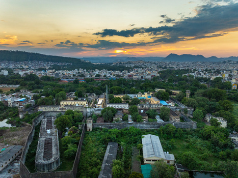 Aerial view of a historic fortified complex surrounded by lush greenery and urban landscape under a golden sunset sky in Udaipur Rajasthan India.
