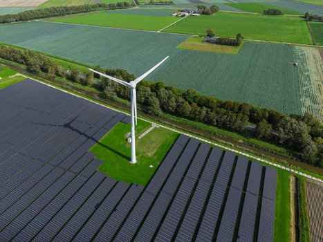 Aerial view of a wind turbine standing amidst a large solar farm surrounded by green agricultural fields and canals in Warmenhuizen North Holland Netherlands.
