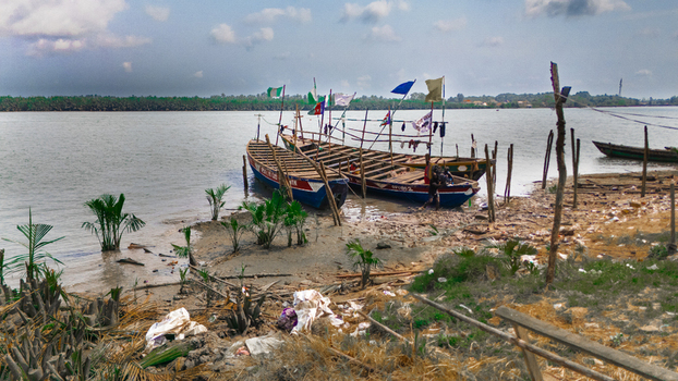 Aerial view of wooden boats moored on a muddy riverbank with flags palm trees and scattered debris along the shoreline in Rivers Nigeria.