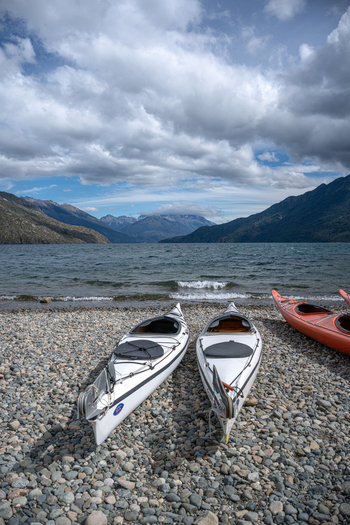 View of two white kayaks resting on the pebble shore of Lago Puelo National Park with mountains and a cloudy sky in the background in Cushamen Chubut Argentina.