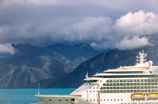 View of a large white cruise ship docked in front of dramatic cloud covered mountains and turquoise water in Haines Alaska United States. View of a large white cruise ship docked in front of dramatic cloud covered mountains and turquoise water in Haines Alaska United States.