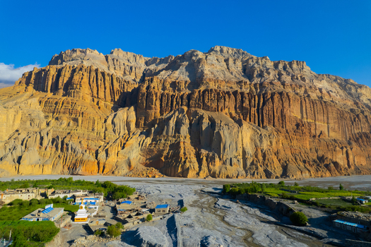 Aerial view of a traditional stupa nestled in a village at the base of towering rugged Himalayan cliffs under a clear blue sky in Nepal.