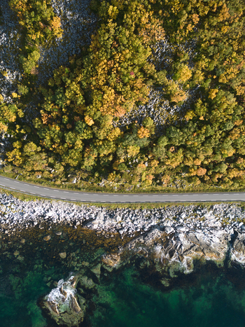 Aerial view of a winding coastal road passing through an autumn forest with yellow trees and a rocky shoreline by turquoise water in Leknes Nordland Norway. Aerial view of a winding coastal road passing through an autumn forest with yellow trees and a rocky shoreline by turquoise water in Leknes Nordland Norway.