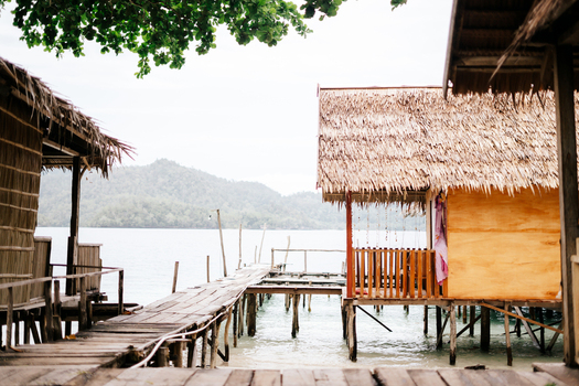 View of traditional wooden stilt houses with thatched roofs connected by a wooden walkway over clear turquoise water in Raja Ampat island Southwest Papua Indonesia. View of traditional wooden stilt houses with thatched roofs connected by a wooden walkway over clear turquoise water in Raja Ampat island Southwest Papua Indonesia.