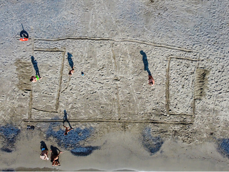 Aerial view of a soccer field drawn in the sand with people playing and long shadows on the beach in San Lorenzo Liguria Italy.